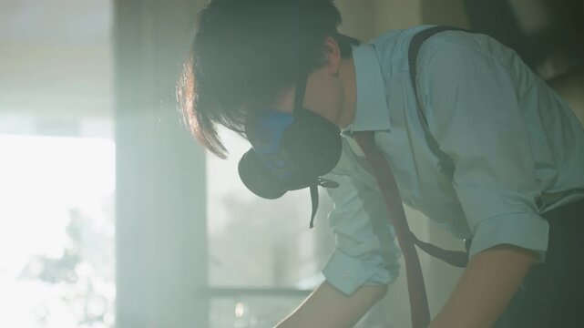 Asian investigator in respirator leans over inspecting tabletop documents, adjusting tools with gloved hands, morning backlight silhouette, cautious posture, protective gear and sterile investigation