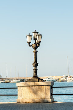Ornate cast iron street lamp with four lanterns on waterfront promenade in Bari, Italy, with marina and sailboats visible in background on clear sunny day