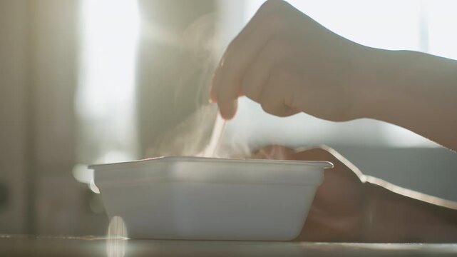 Sunlit closeup hand stirring steaming meal, disposable white container on tabletop, plastic spoon lifting hot rice and sauce, gentle steam rising through warm backlight, home kitchen window glow,