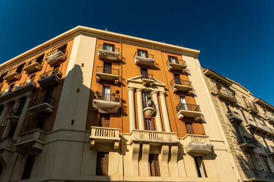 Historic apartment building facade in Bari, Italy, with classical columns, iron balconies and warm orange render under deep blue sky on sunny day