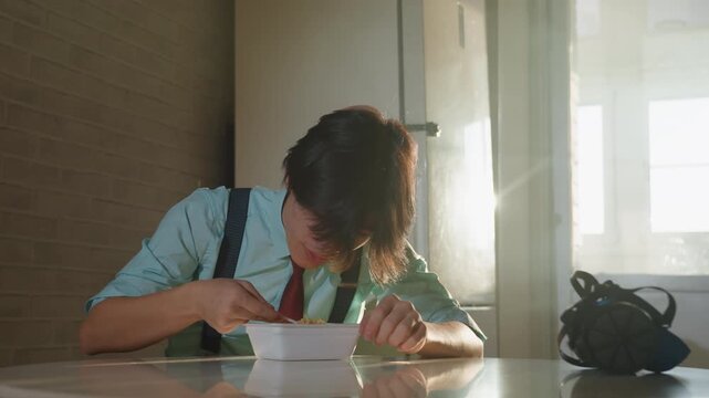 Backlit young man eating noodles at table, sunlight flooding through window, backpack at side, slow focused movements, silent contemplative mood, office interior with warm cinematic tones, tie