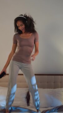 Happy African American woman listening music in headphones having fun jumping joy on bed at evening. Joyous female dance jump fooling around in bedroom girl listen song with mobile phone earphones