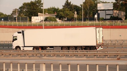 Biały samochód ciężarowy z naczepą chłodniczą jedzie drogą szybkiego ruchu wzdłuż ekranu akustycznego. Zdjęcie przedstawia profil pojazdu realizującego transport towarowy i logistyczny.
