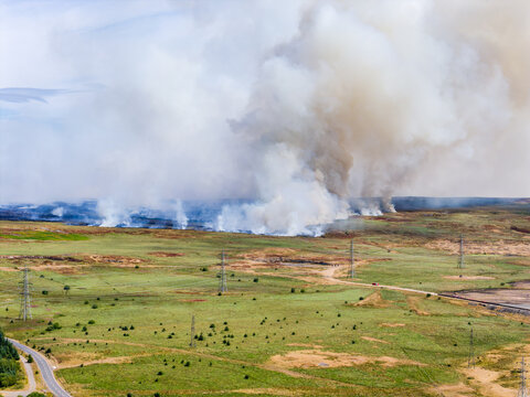 Thick smoke plume rising from grassland wildfire in open countryside, fire line spreading across dry hills past power pylons and countryside road under summer sky