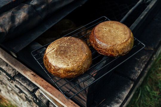 Two wheels of homemade smoked goat cheese resting on a metal rack inside a rustic outdoor smoker, showing a dark golden rind and traditional artisanal preparation