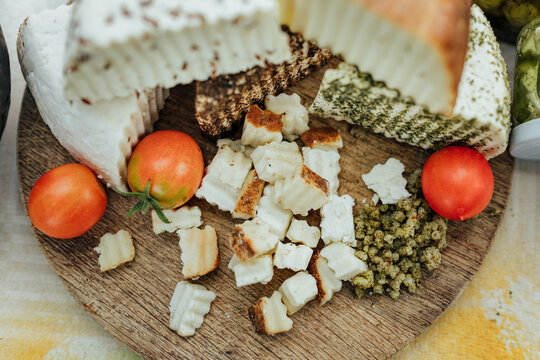 Assorted fresh, herb-coated and aged goat cheeses on a wooden board with tomatoes, creamy spread and fruit preserve, presented as a rustic gourmet tasting
