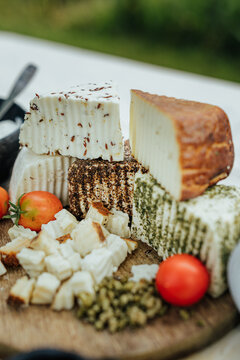 Assorted fresh, herb-coated and aged goat cheeses on a wooden board with tomatoes, creamy spread and fruit preserve, presented as a rustic gourmet tasting