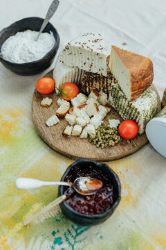 Assorted fresh, herb-coated and aged goat cheeses on a wooden board with tomatoes, creamy spread and fruit preserve, presented as a rustic gourmet tasting