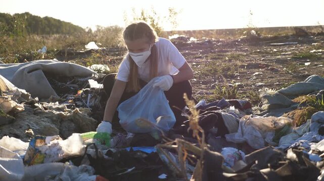 Close up of little girl in protective mask collecting waste in bag near roadside. Small female eco volunteer in gloves cleaning lawn of trash at countryside. Concept of environmental pollution problem