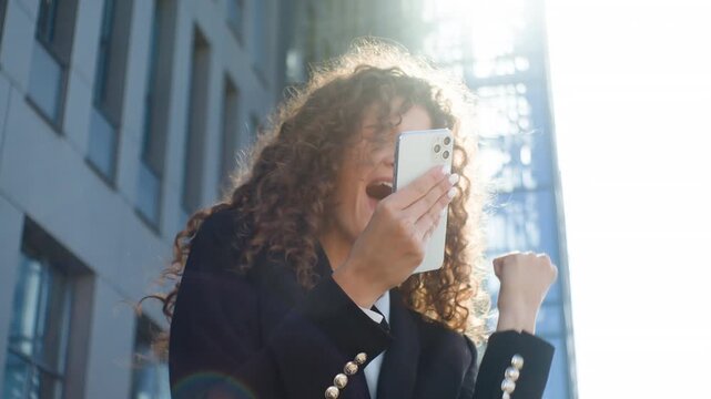 Emotional positive Caucasian business woman winning with mobile phone dancing happy in city European businesswoman girl female agent expert user win with smartphone dance ecstatic in sun light in city