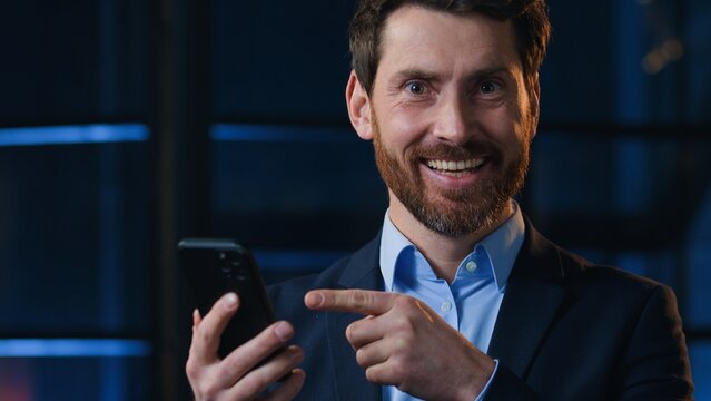 Caucasian middle-aged smiling business man pointing at mobile phone at night evening office mature businessman boss male executive holding smartphone excited with online news message looking at camera