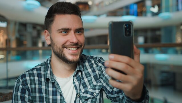 Happy smiling Caucasian man influencer talking video call with mobile phone in shopping mall cheerful male blogger talk chatting online conference with smartphone recording blog social media stream