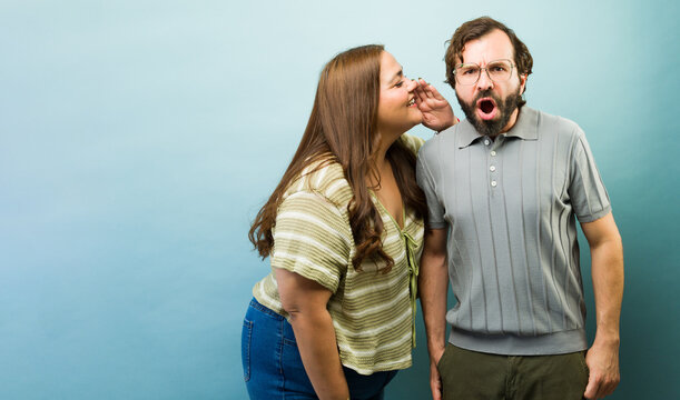 Curvy woman whispering a secret into man's ear, causing a shocked and surprised reaction, sharing gossip and rumors