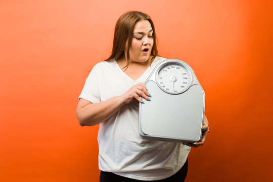 Plus size young woman holding a weight scale, feeling surprised and concerned about her body weight and health