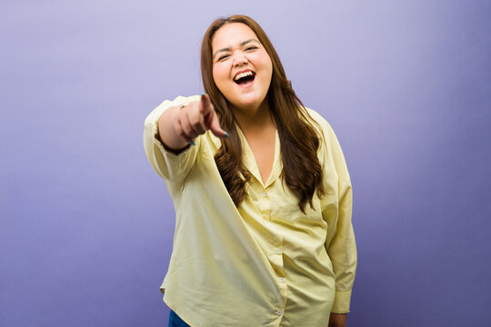 Happy young plus size woman pointing finger, laughing and smiling at camera, feeling joy and amusement on purple background