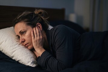 Obraz premium A woman is in bed during the night. She has her hands on her face and looks lost in thought. The room is dimly lit and she appears to be awake and reflecting