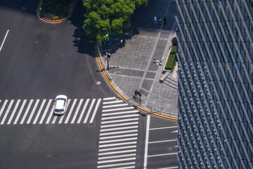 Overhead view of a rounded office forecourt where crossings meet a busy carriageway, a white car easing across the striped crossing toward the corner, in the core of a busy business quarter. © ultramansk