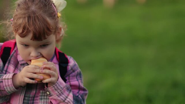 Child girl with backpack holding a pastry while walking in the park. Caucasian kid eating a bun during a sunny day. Outdoor lifestyle and childhood concept.