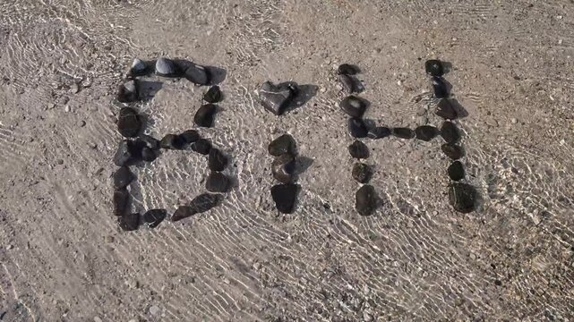 Stones arranged to spell BIH on a river bank splashed by water. Pebbles spelling the abbreviation for Bosnia and Herzegovina on the shore of a flowing river.