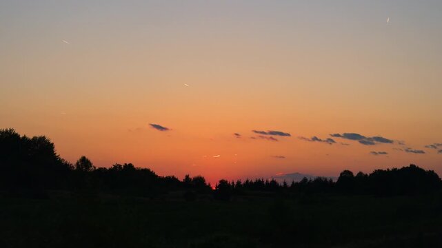 Vibrant sunset or sunrise with a warm glowing orange and red sky above a dark treeline silhouette of trees on a mountain. Colorful twilight sky with dramatic orange and red tones.