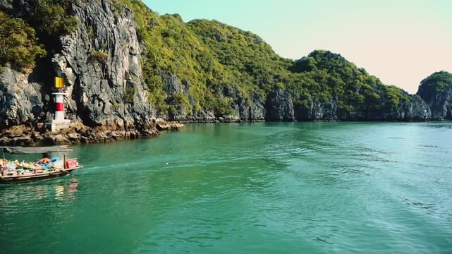 A motorized boat carrying tourists navigates the calm emerald waters of Ha Long Bay, Vietnam, surrounded by towering limestone cliffs covered in lush green vegetation during daytime.