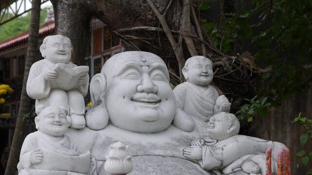 White laughing budai statue in a tropical garden with small buddha figures and greenery in the background. Good fortune.