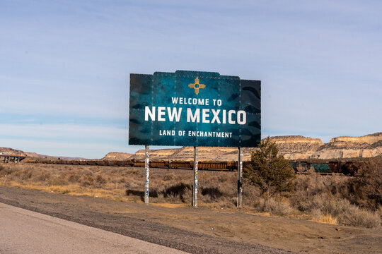 Welcome to New Mexico sign featuring the Zia sun symbol and &ldquo;Land of Enchantment,&rdquo; with desert mesas, sparse vegetation, railroad tracks, and a passing train