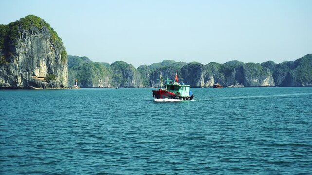 A motorized tourist boat cruises across the deep blue waters of Halong Bay, surrounded by towering limestone karst formations covered in lush green vegetation in Vietnam.