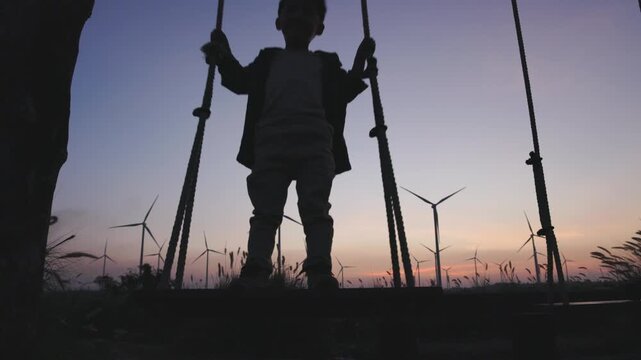 Silhouette of a young boy standing on a swing at sunset with wind turbines in the background. Peaceful scene of a child enjoying nature and clean renewable energy during golden hour outdoors