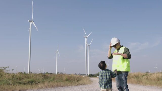 Asian engineer father putting a safety helmet on his young son at a wind turbine farm. Inspiring moment of family bonding and learning about green renewable energy outdoors
