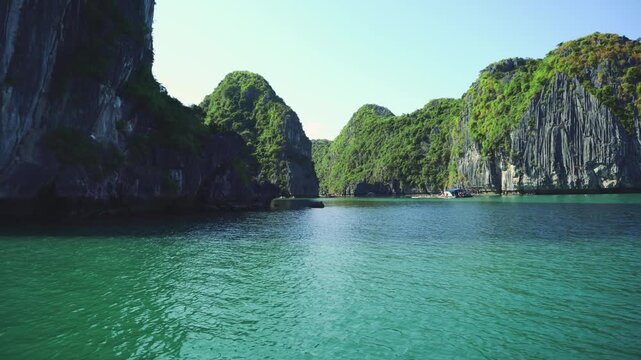 A panoramic daytime view of turquoise water and steep limestone karsts covered with dense tropical foliage in Halong Bay, Vietnam, with small boats near the islands.