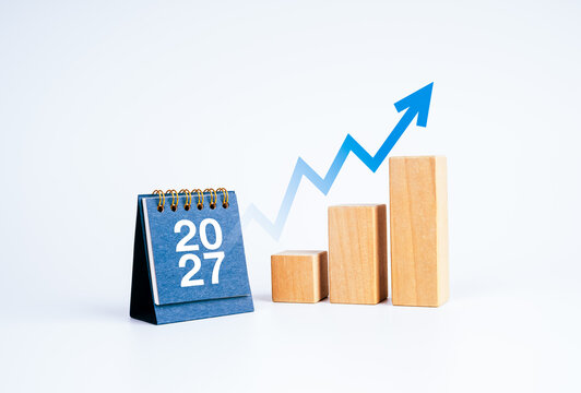 Blue 2027 desk calendar beside rising wooden bars graph steps and an upward arrow on clean white background. Business growth, financial forecasting, strategic planning and future success concept.