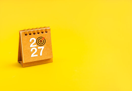 Desk calendar displays 2027 with bullseye replacing zero on bright yellow background with copy space. Annual goals, deadline management, strategic planning, focus and measurable achievement concept.