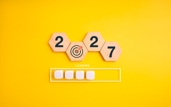 Wooden hexagon blocks form 2027 with bullseye replacing zero above loading bar on a yellow background. Preparation, progress tracking, strategic goals and readiness for the new year concept.