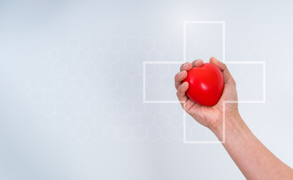 Hand holding a red heart ball with medical cross symbol representing heart health, healthcare, preventive care, cardiology, patient care, wellness, medical services, and health insurance awareness.