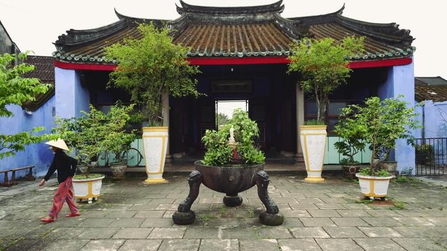Two people walk through a traditional temple courtyard with blue walls and tiled roof in Hoi An, Vietnam at nighttime, surrounded by stone planters and greenery.