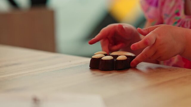 Girl touching chocolate flower cookie on wooden table. Child playing with biscuit dessert. Snack time activity, sweet food interaction, bakery product enjoyment.