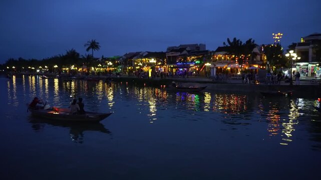 People carrying illuminated lanterns on boats along the river at night in Hoi An, Vietnam, with brightly lit historic buildings and palm trees lining the waterfront promenade.