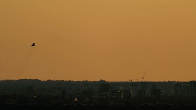 Airplane climbs into the sky above the dark buildings of Perth at sunset with golden light filling the horizon during evening