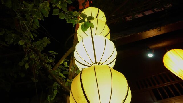 A stack of five large illuminated paper lanterns glowing warmly at night in the ancient town of Hoi An, Vietnam, framed by green foliage and traditional architecture.