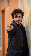 Fototapeta premium A confident young man with curly dark hair and facial hair wearing a black jacket points directly at camera while leaning against orange wall, creating engaging portrait.