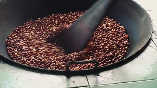 A close-up of peeled peanuts being stirred with a dark paddle inside a large metal pan on a light indoor surface. No people or animals are visible.