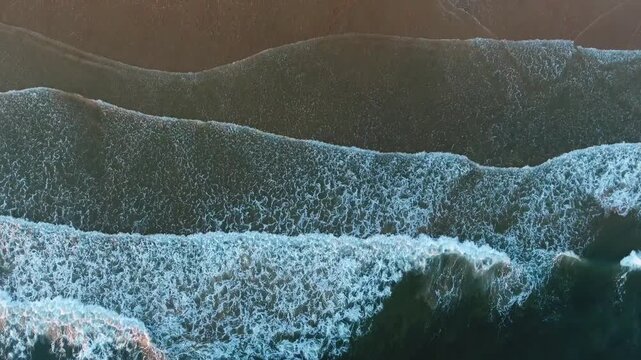 Majestic aerial top down view of powerful turquoise ocean waves rolling and crashing onto a serene, sandy shoreline, creating a beautiful and mesmerizing natural pattern on the water's surface