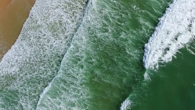 Stunning aerial top down perspective of beautiful green ocean waves with white foam rolling onto a sandy shore, creating a mesmerizing and relaxing natural pattern on the water surface