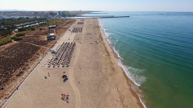 Aerial view of the expansive sandy beach of vilamoura in portugal, showing the turquoise ocean water, beach umbrellas, and the city skyline in the distance on a sunny summer day