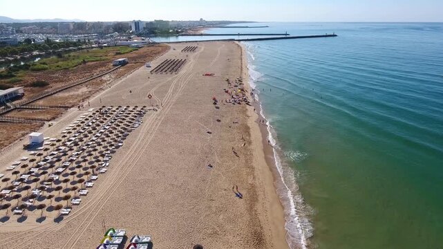Aerial shot flying over the beautiful sandy beach of vilamoura in the algarve region, showing tourists relaxing under umbrellas next to the turquoise sea and the city skyline in the background