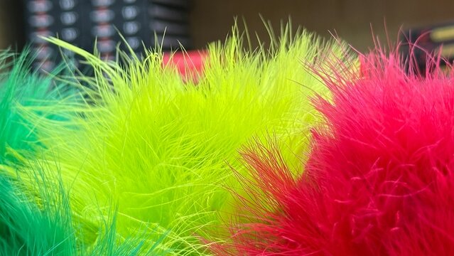 A vibrant close-up macro shot featuring fluffy, brightly dyed green, yellow, and red feathers or long-pile faux fur creating a soft, colorful, and playful abstract texture.