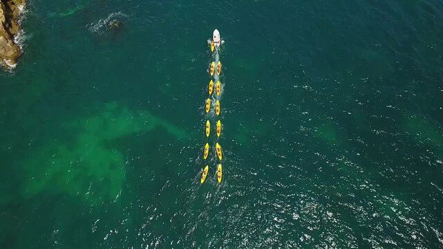 Aerial top down view shows a group of tourists on yellow kayaks following a guide on a boat, paddling in formation through the beautiful turquoise ocean water during a summer excursion