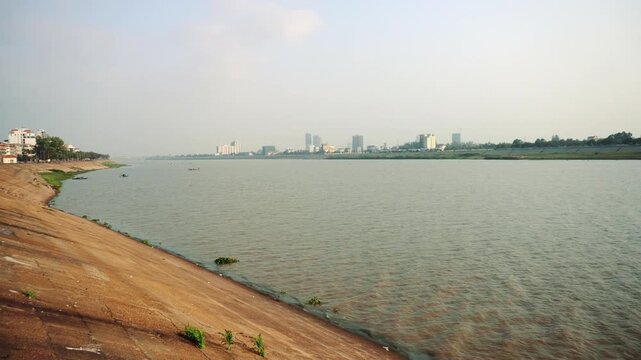 A wide-angle daytime view of the Mekong River flowing alongside Phnom Penh's urban embankment with modern buildings and a small boat in the distance.