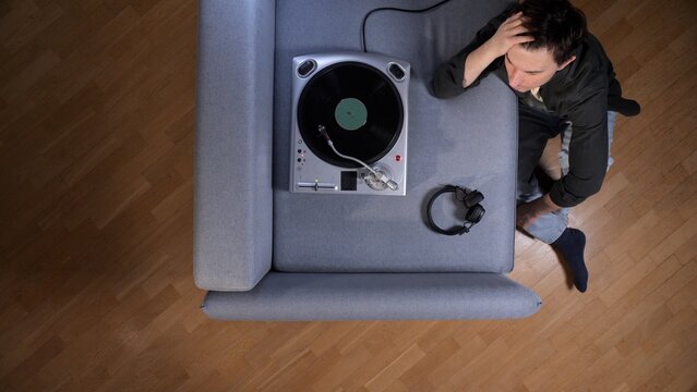 Man sitting on the floor by a turntable and couch, listening to vinyl records through headphones, reflecting on loneliness and melancholy